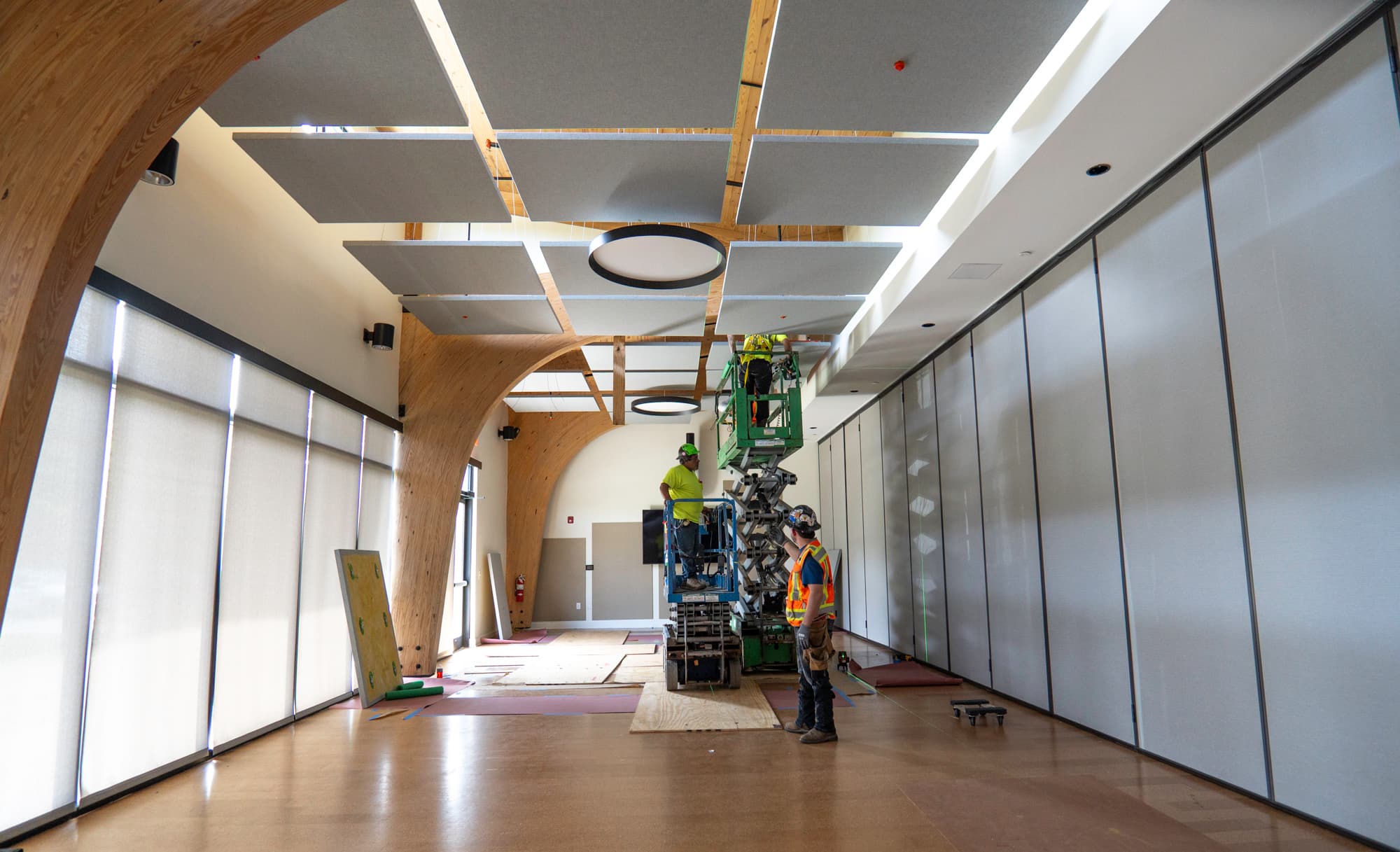 Crew installing acoustical ceiling clouds and specialty panels beneath a curved timber structure in a daylit interior